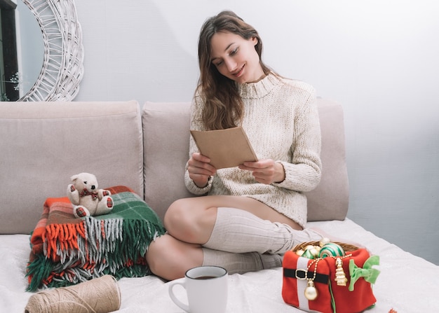 A young pretty woman in a light sweater sits on a bed with Christmas decorations and reads a letter