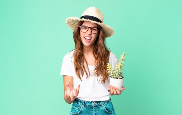 Young pretty woman looking angry, annoyed and frustrated with a straw hat and holding a cactus