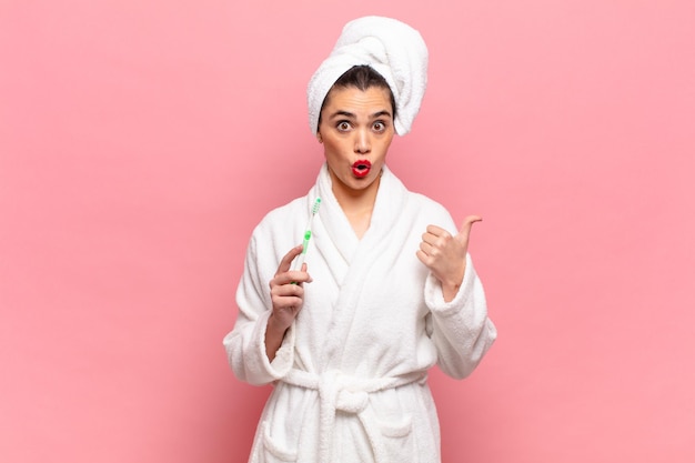 Young pretty woman looking astonished in disbelief, pointing at object on the side and saying wow, unbelievable. bathrobe and toothbrush concept