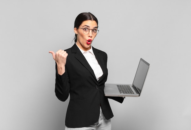 Young pretty woman looking astonished in disbelief, pointing at object on the side and saying wow, unbelievable. laptop concept