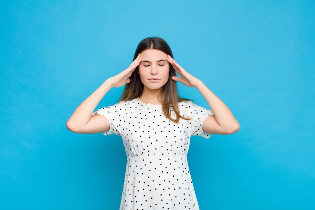 Young pretty woman looking concentrated, thoughtful and inspired, brainstorming and imagining with hands on forehead against blue wall