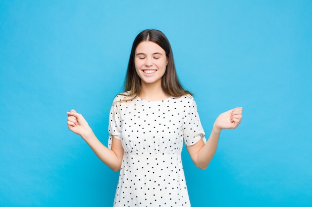 Young pretty woman looking extremely happy and surprised, celebrating success, shouting and jumping over blue wall