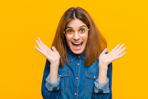 Young pretty woman looking happy and excited, shocked with an unexpected surprise with both hands open next to face against yellow background