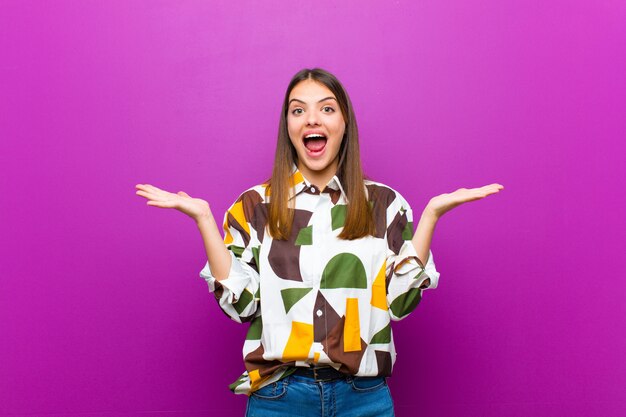 Young pretty woman looking happy and excited, shocked with an unexpected surprise with both hands open next to face over purple wall