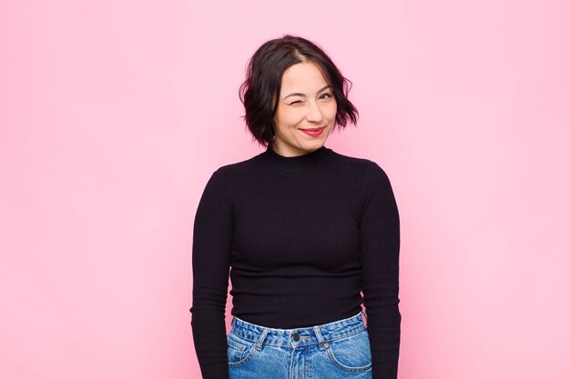 Young pretty woman looking happy and friendly, smiling and winking an eye at you with a positive attitude against pink wall