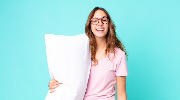 Молодая красивая женщина выглядит счастливой и приятно удивленной в пижаме и держит подушку