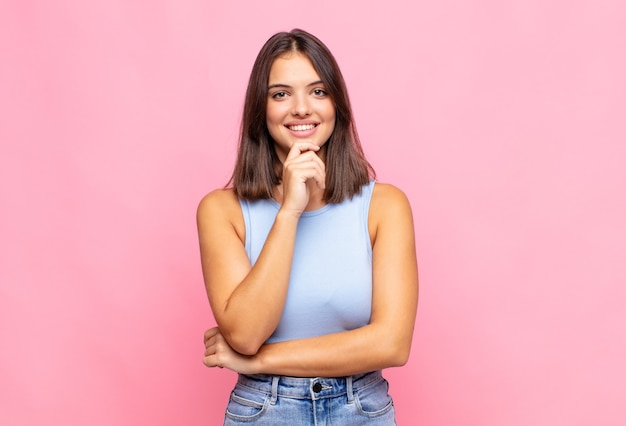 Giovane donna graziosa che sembra felice e sorridente con la mano sul mento, chiedendosi o facendo una domanda, confrontando le opzioni