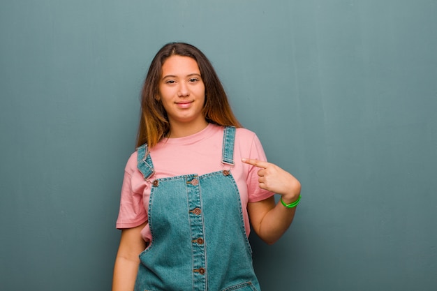 Young pretty  woman looking proud, confident and happy, smiling and pointing to self or making number one sign against grunge wall