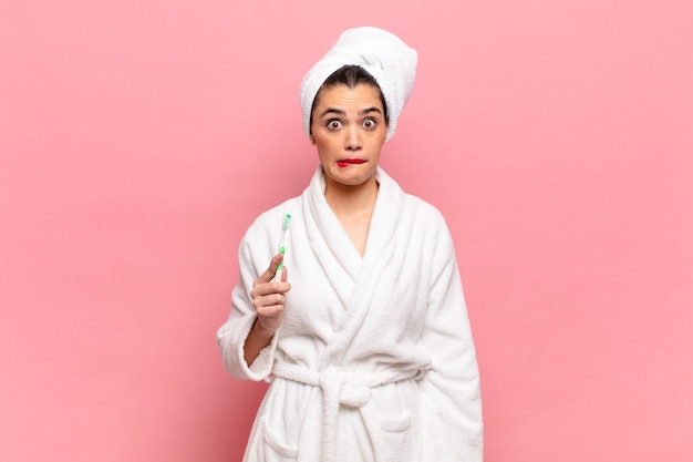 Young pretty woman looking puzzled and confused, biting lip with a nervous gesture, not knowing the answer to the problem. bathrobe and toothbrush concept
