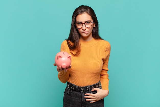 Young pretty woman looking puzzled and confused, biting lip with a nervous gesture, not knowing the answer to the problem. piggy bank concept