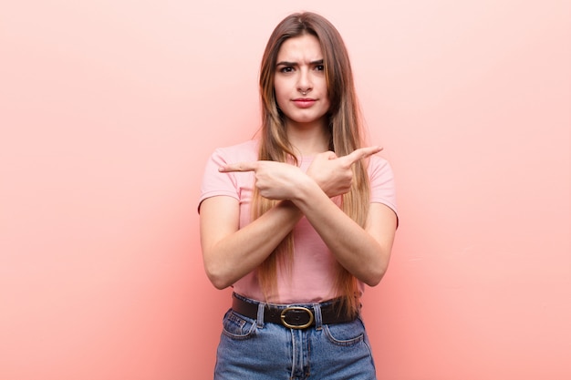 Foto giovane donna graziosa che sembra perplessa e confusa, insicura e che punta in direzioni opposte con dubbi contro il muro rosa