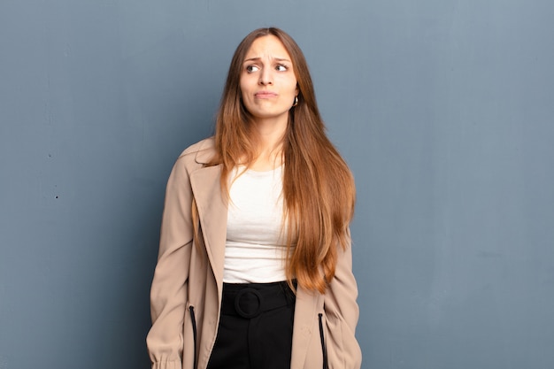 Young pretty woman looking puzzled and confused, wondering or trying to solve a problem or thinking over gray wall