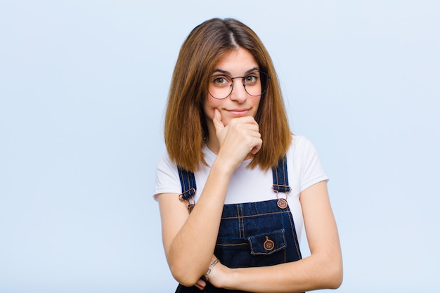 Young pretty woman looking serious, confused, uncertain and thoughtful, doubting among options or choices against blue wall