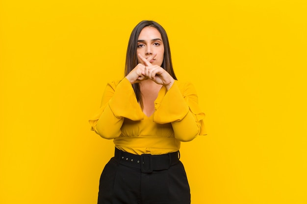 Young pretty woman looking serious and displeased with both fingers crossed up front in rejection, asking for silence over orange wall