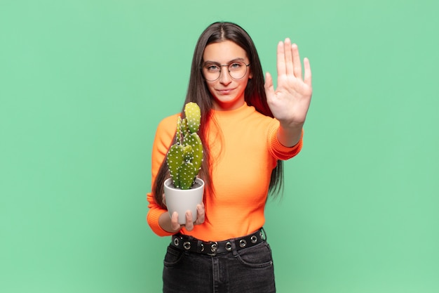 Giovane bella donna che sembra seria, severa, dispiaciuta e arrabbiata che mostra il palmo aperto che fa il gesto di arresto. concetto di cactus