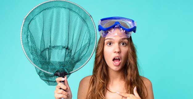 写真 ゴーグルと漁網で自分を指差しながら、口を大きく開けてショックを受けて驚いた若いきれいな女性