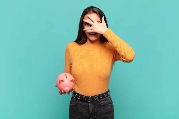 Young pretty woman looking shocked, scared or terrified, covering face with hand and peeking between fingers. piggy bank concept