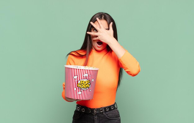 Young pretty woman looking shocked, scared or terrified, covering face with hand and peeking between fingers. pop corns concept