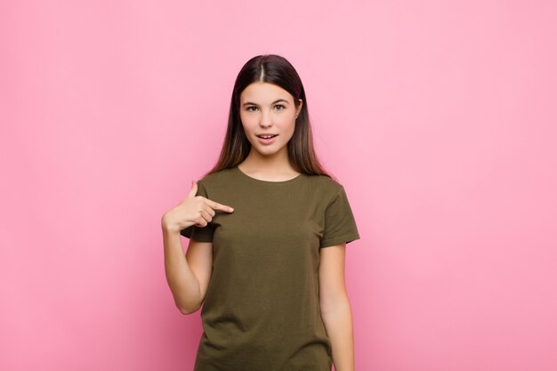 Young pretty woman looking shocked and surprised with mouth wide open, pointing to self against pink wall