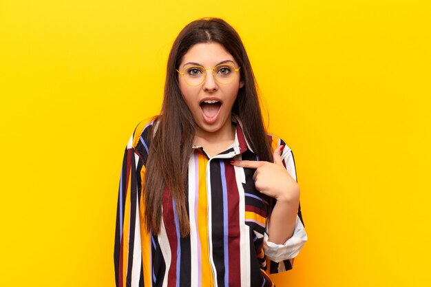 Young pretty woman looking shocked and surprised with mouth wide open, pointing to self against yellow wall