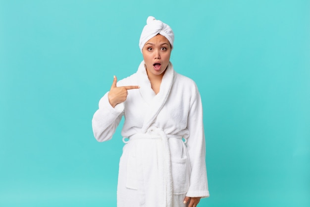 Young pretty woman looking shocked and surprised with mouth wide open, pointing to self and wearing bath robe after shower