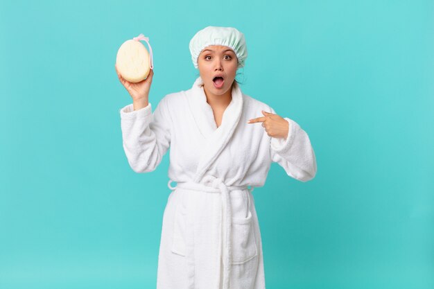 Young pretty woman looking shocked and surprised with mouth wide open, pointing to self and wearing bath robe after shower