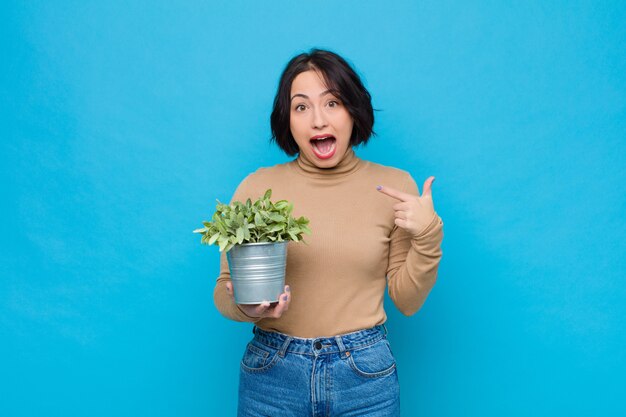 Young pretty woman looking shocked and surprised with mouth wide open, pointing to self with a plant