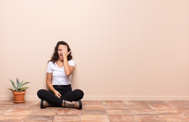 Young pretty woman looking sleepy, bored and yawning, with a headache and one hand covering half the face sitting a terrace floor