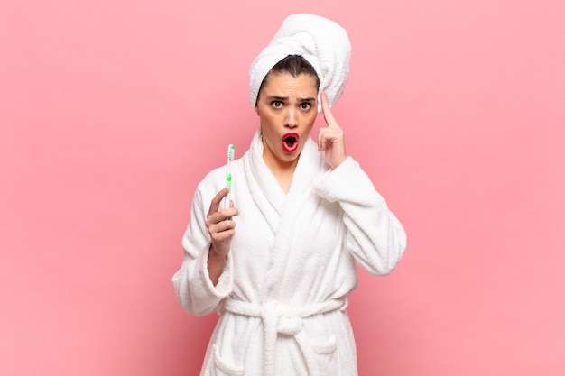 Young pretty woman looking surprised, open-mouthed, shocked, realizing a new thought, idea or concept. bathrobe and toothbrush concept