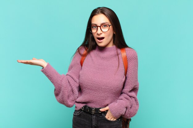 Giovane donna graziosa che sembra sorpresa e scioccata, con la mascella caduta tenendo un oggetto con una mano aperta sul lato. concetto di studente