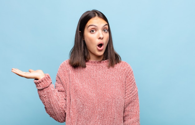 Young pretty woman looking surprised and shocked, with jaw dropped holding an object with an open hand on the side