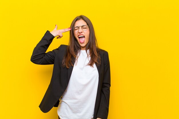 Young  pretty woman looking unhappy and stressed, suicide gesture making gun sign with hand, pointing to head against orange wall