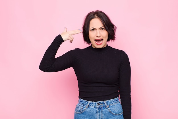 Young pretty woman looking unhappy and stressed, suicide gesture making gun sign with hand, pointing to head against pink wall