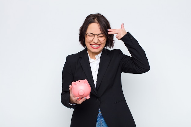 Young pretty woman looking unhappy and stressed, suicide gesture making gun sign with hand, pointing to head with a piggy bank