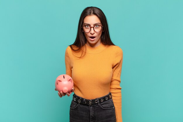 Young pretty woman looking very shocked or surprised, staring with open mouth saying wow. piggy bank concept