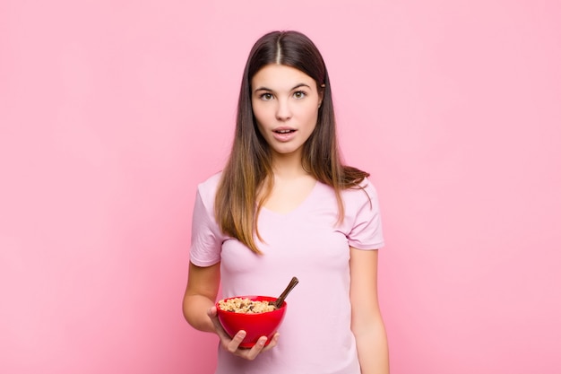 Young pretty woman looking very shocked or surprised, staring with open mouth saying wow with a breakfast bowl