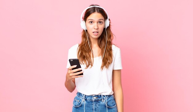 Young pretty woman looking very shocked or surprised with headphones and a smartphone