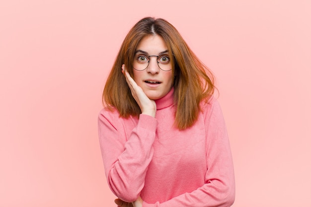 Young pretty woman open-mouthed in shock and disbelief, with hand on cheek and arm crossed, feeling stupefied and amazed against pink wall