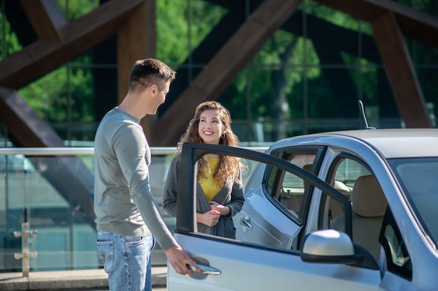 Young pretty woman opening the car door, talking to the young man and smiling