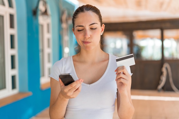 屋外で考えながらクレジット カードで携帯電話で購入する若いきれいな女性