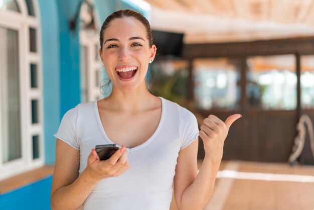 Young pretty woman at outdoors using mobile phone and pointing to the lateral