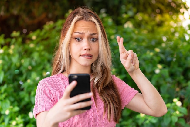 Young pretty woman at outdoors using mobile phone with fingers crossing