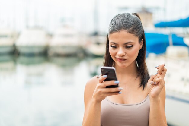 Young pretty woman at outdoors using mobile phone with fingers crossing