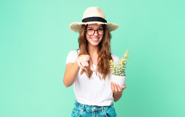 Young pretty woman pointing at camera choosing you with a straw hat and holding a cactus
