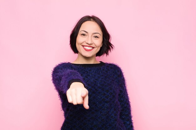 Young pretty woman pointing at camera with a satisfied, confident, friendly smile, choosing you over pink wall