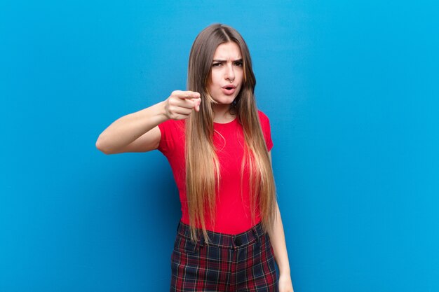 Young pretty woman pointing with an angry aggressive expression looking like a furious, crazy boss against blue wall