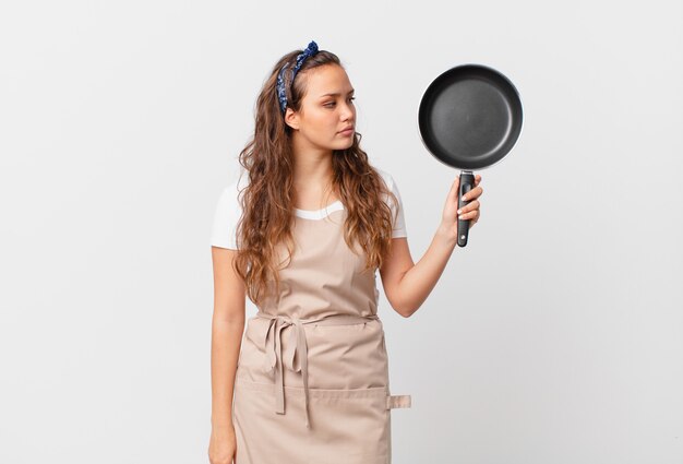 Giovane bella donna in vista di profilo pensando, immaginando o sognando ad occhi aperti il concetto di chef e tenendo in mano una padella