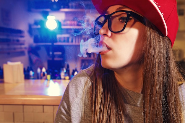 Giovane donna graziosa in berretto rosso fuma una sigaretta elettronica al negozio di vape primo piano