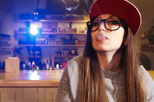 Young pretty woman in red cap smoke an electronic cigarette at the vape shop. Closeup.