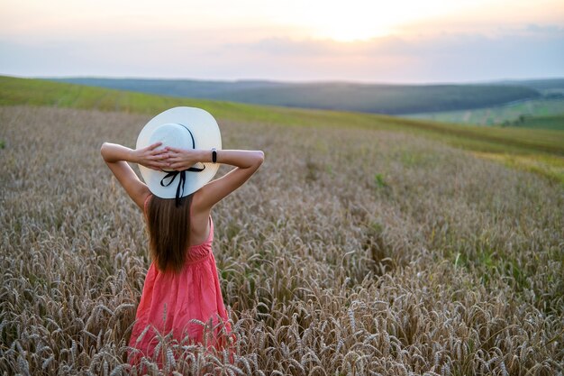 赤い夏のドレスと黄色の農地に立っている麦わら帽子の若いきれいな女性